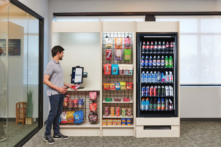 A person selects snacks from a modern office vending station with a variety of chips, candy, and drinks alongside a